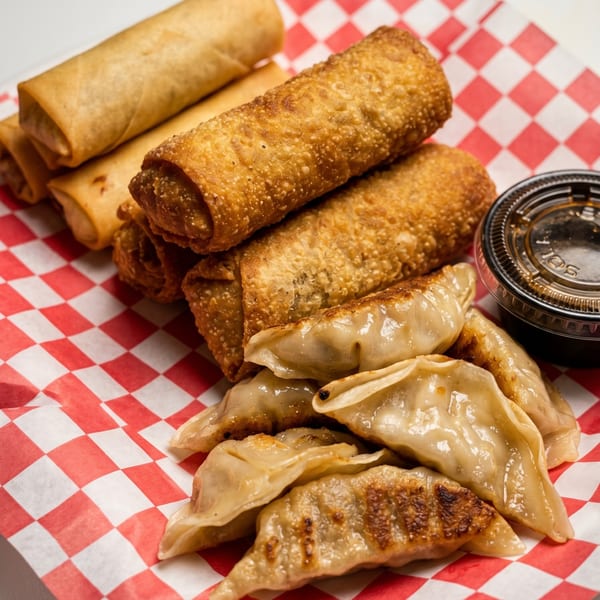Crispy egg rolls and pan-fried dumplings with dipping sauce on red and white checkered paper.