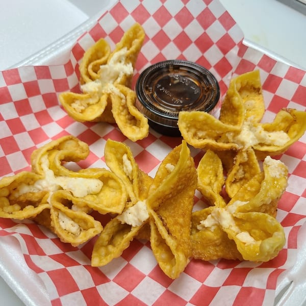 Golden-fried crab rangoons with creamy filling and a side of dipping sauce on red and white checkered paper.